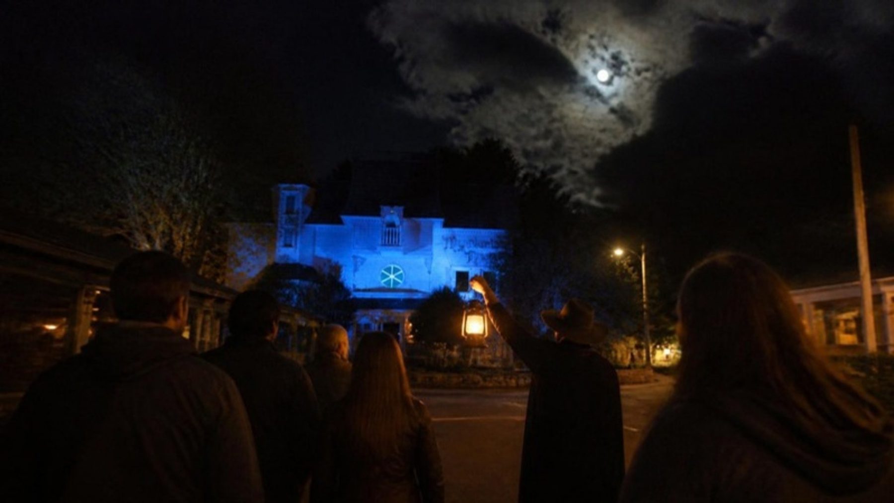 A small evening tour group seen from behind on a dark street, facing a historic house lit in blue under a full moon