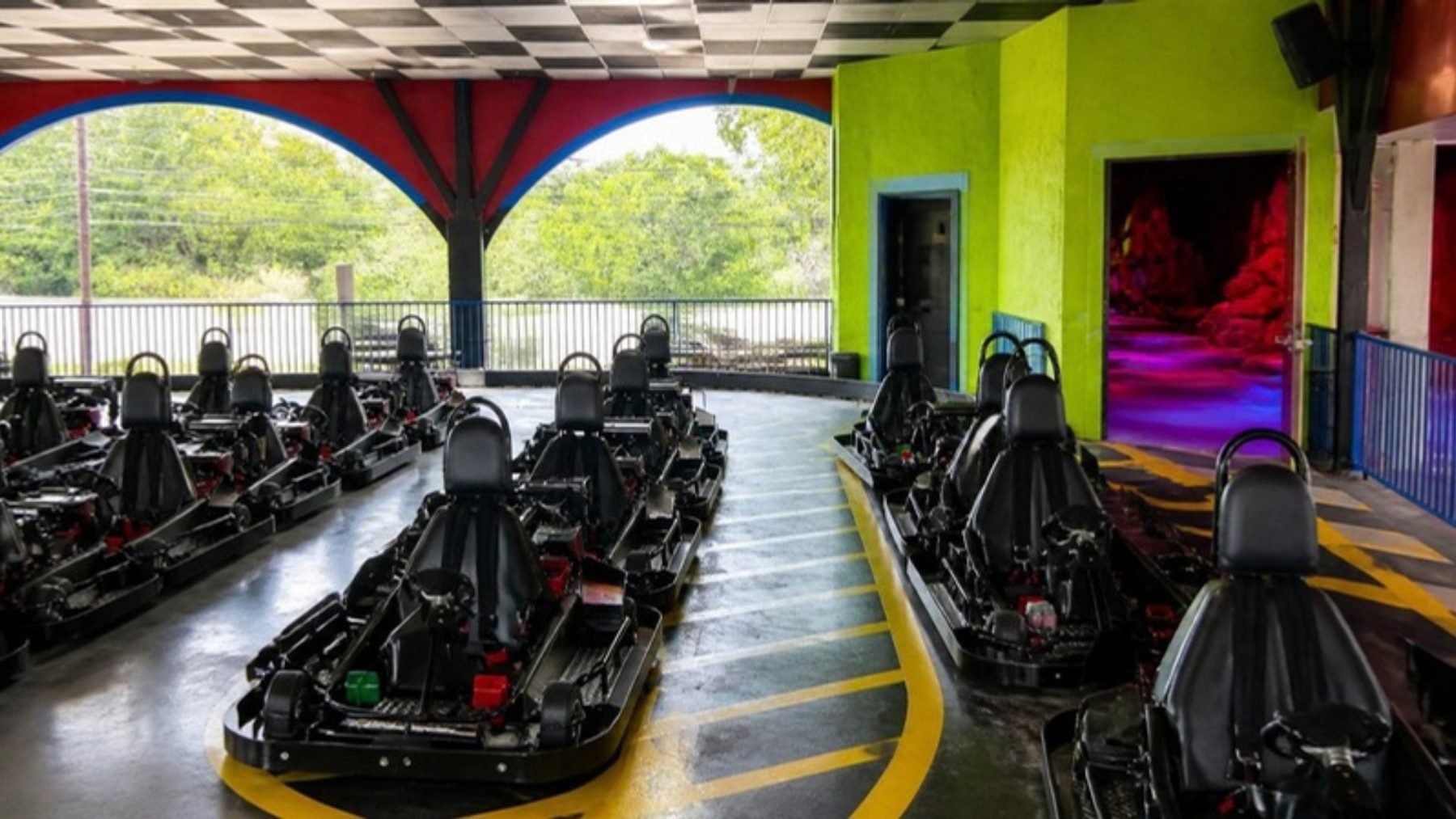 Rows of go-karts lined up on a covered slick track with yellow edge striping and lime-green walls, a neon-lit doorway beyond
