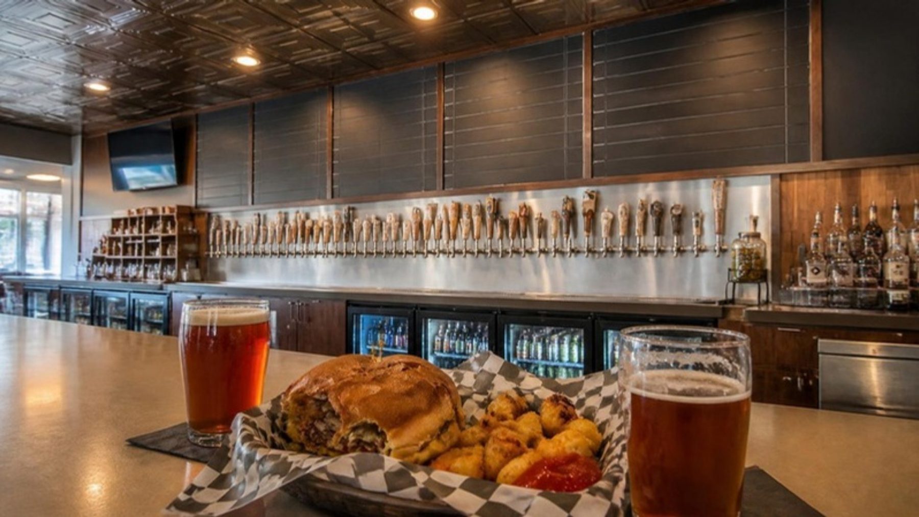 Craft beer bar interior with a long wall of beer taps, pints of amber beer and a burger basket on the bar in the foreground