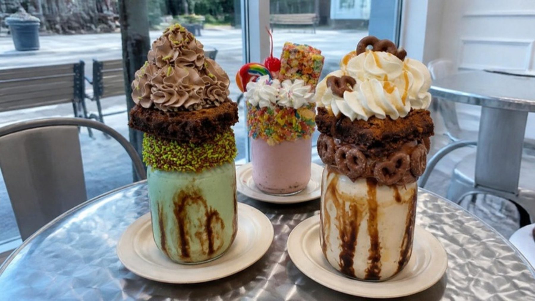 Towering mason-jar milkshakes topped with brownies, whipped cream and candy on a steel table in a bright shake bar