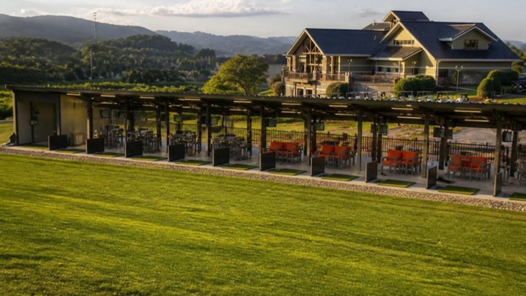 Covered Toptracer driving range pavilion with orange-cushioned lounge bays facing a green range, clubhouse and foothills behind