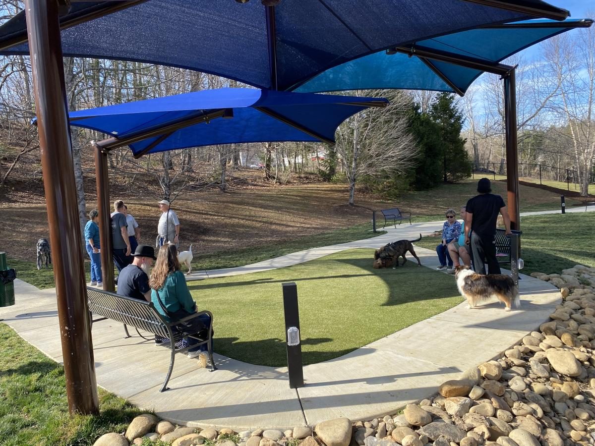 Dogs and owners under blue shade sails on the turf and walking paths of the Gatlinburg Dog Park at Mills Park