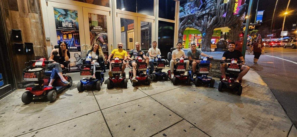 A group of adults seated on rented mobility scooters lined up on a Gatlinburg Parkway sidewalk at night
