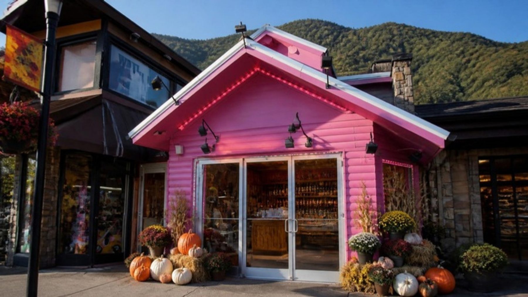 Bright pink gabled winery storefront on the Gatlinburg Parkway with glass doors, pumpkins and mums at the entrance, and a green mountain ridge behind