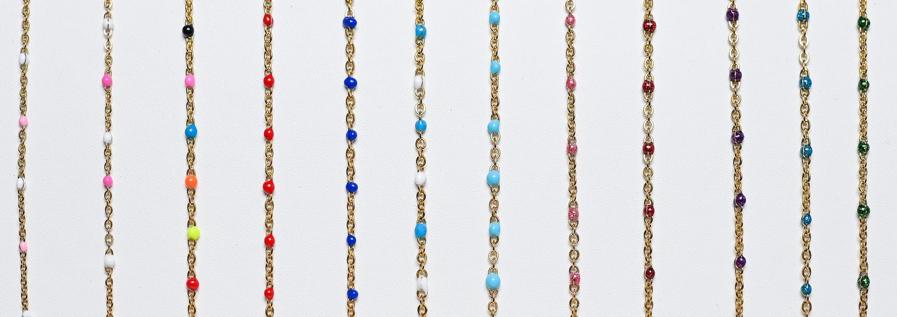 Row of gold permanent-jewelry chains with colorful enamel beads laid out on a white surface