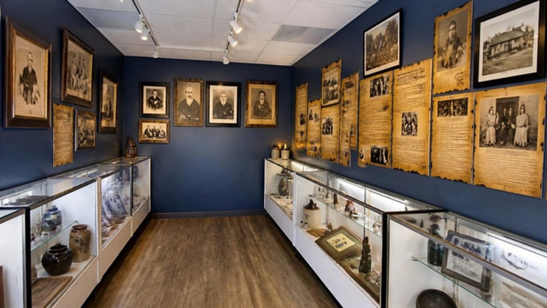 Museum gallery with navy walls lined with framed antique family portraits, history panels and lit glass artifact cases