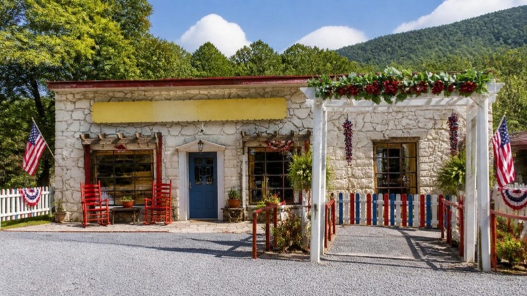 Whitewashed stone general store with a blue door, rocking chairs, a white pergola entry and patriotic bunting below wooded hills