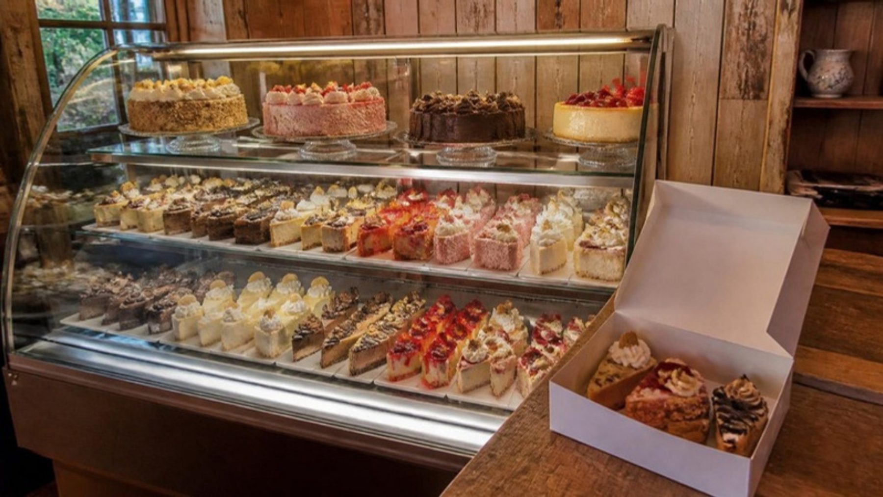 Glass bakery case of assorted cheesecake slices with a takeout box of four slices on a wood counter in a rustic shop