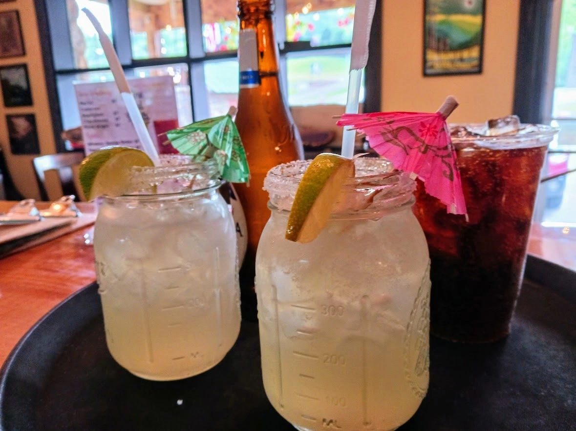 Salt-rimmed margaritas in mason jars and drinks on a tray at a window table inside Little River Pub and Deli