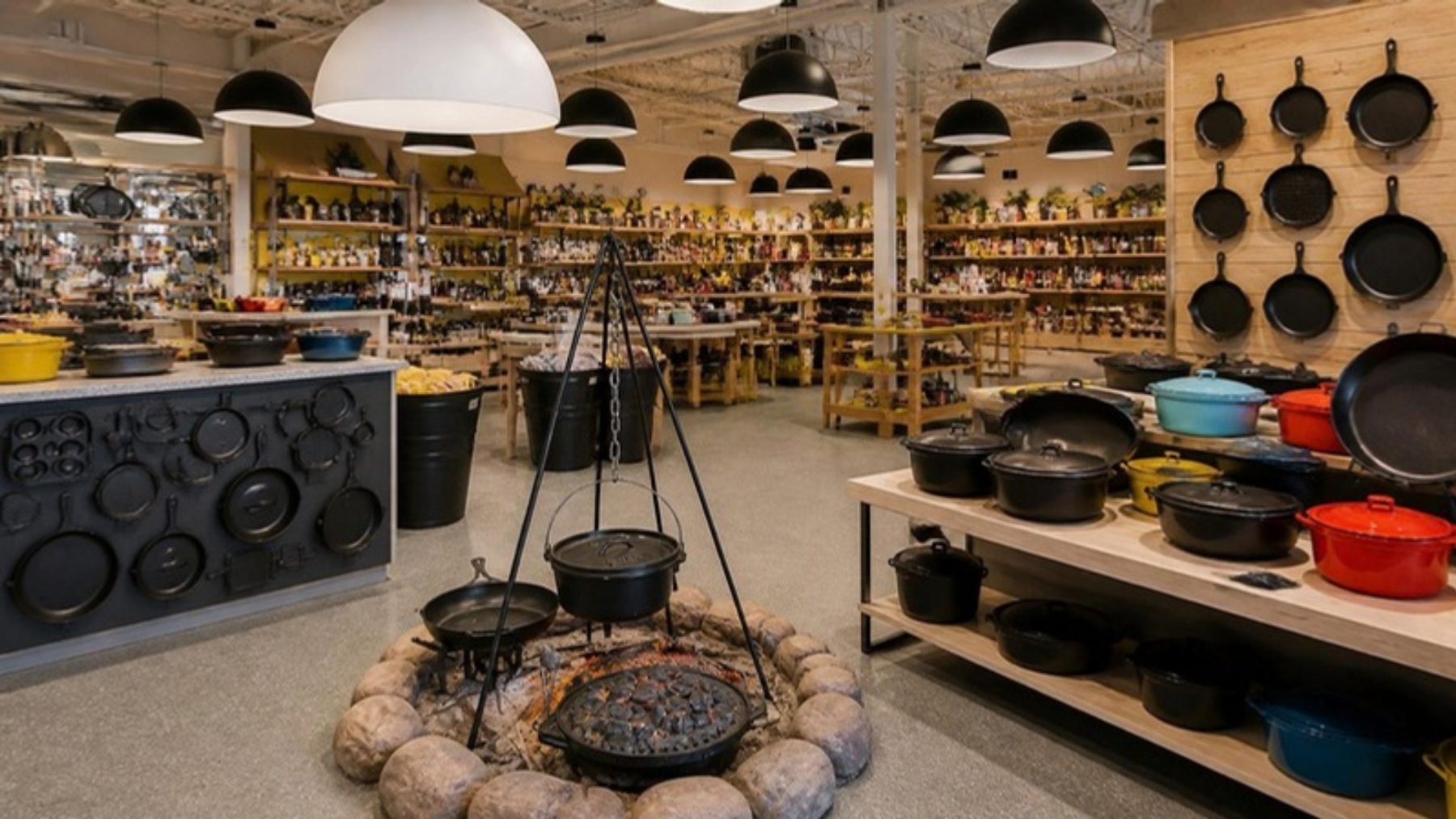 Bright cast iron showroom with dome pendant lights, walls of black skillets and colorful enamel Dutch ovens, and a campfire cooking display in front