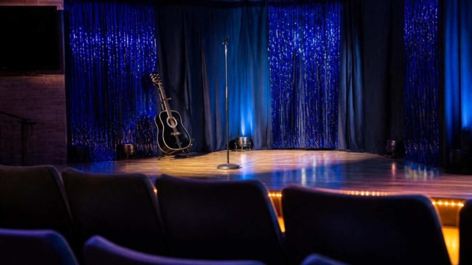 Small theater stage with blue tinsel curtains, an acoustic guitar on a stand and a microphone under stage lights, seen past rows of dark seats