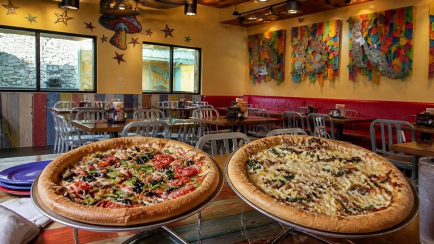 Two stone-baked pizzas on silver trays in a bright dining room with red booths and colorful folk-art murals