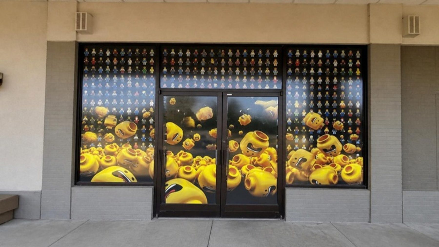 Glass storefront of a minifigure museum with window graphics of rows of tiny figures and giant yellow minifigure heads