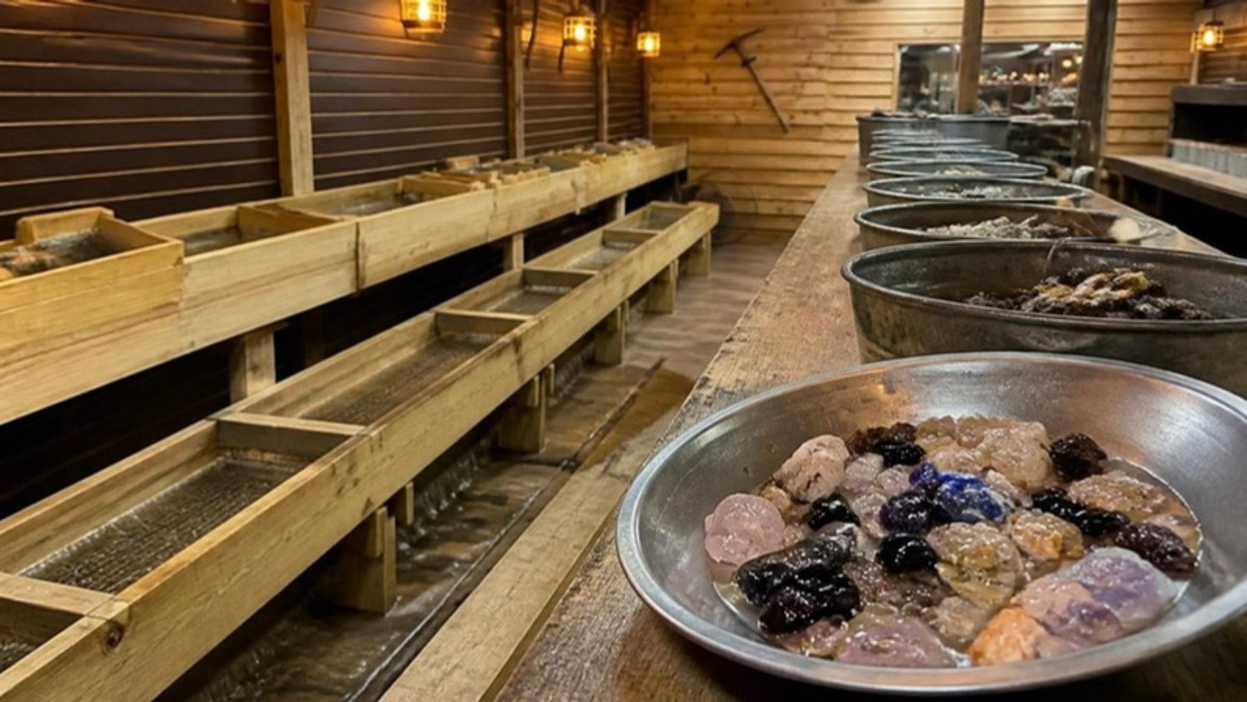 Indoor gem mining room with a long wooden water flume and sifting screens along pine walls, and a metal pan of rough gems and crystals in the foreground
