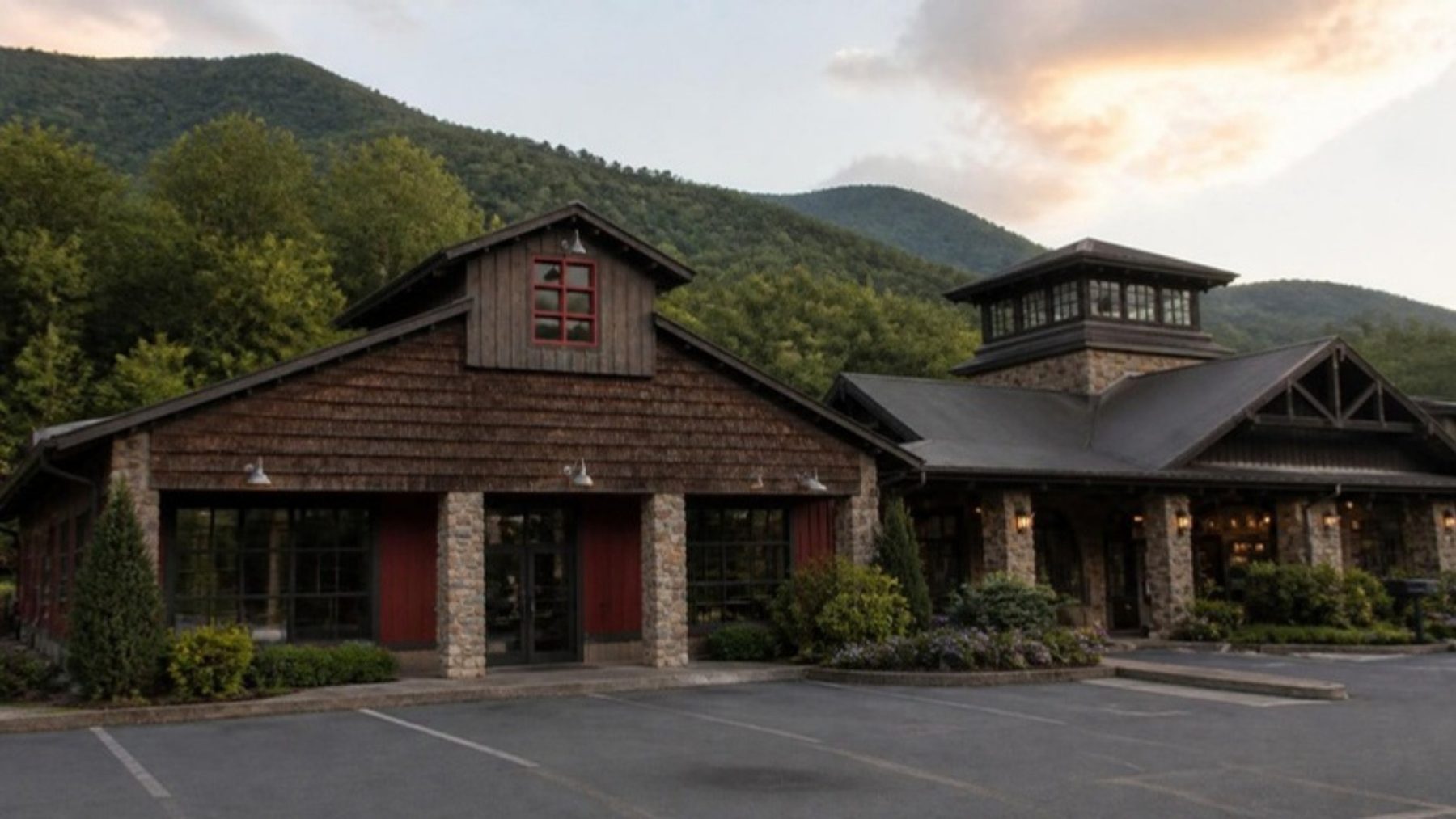 Rustic cedar-shake general store with stone pillars and a gable window beside a stone and timber lodge building, forested mountains behind
