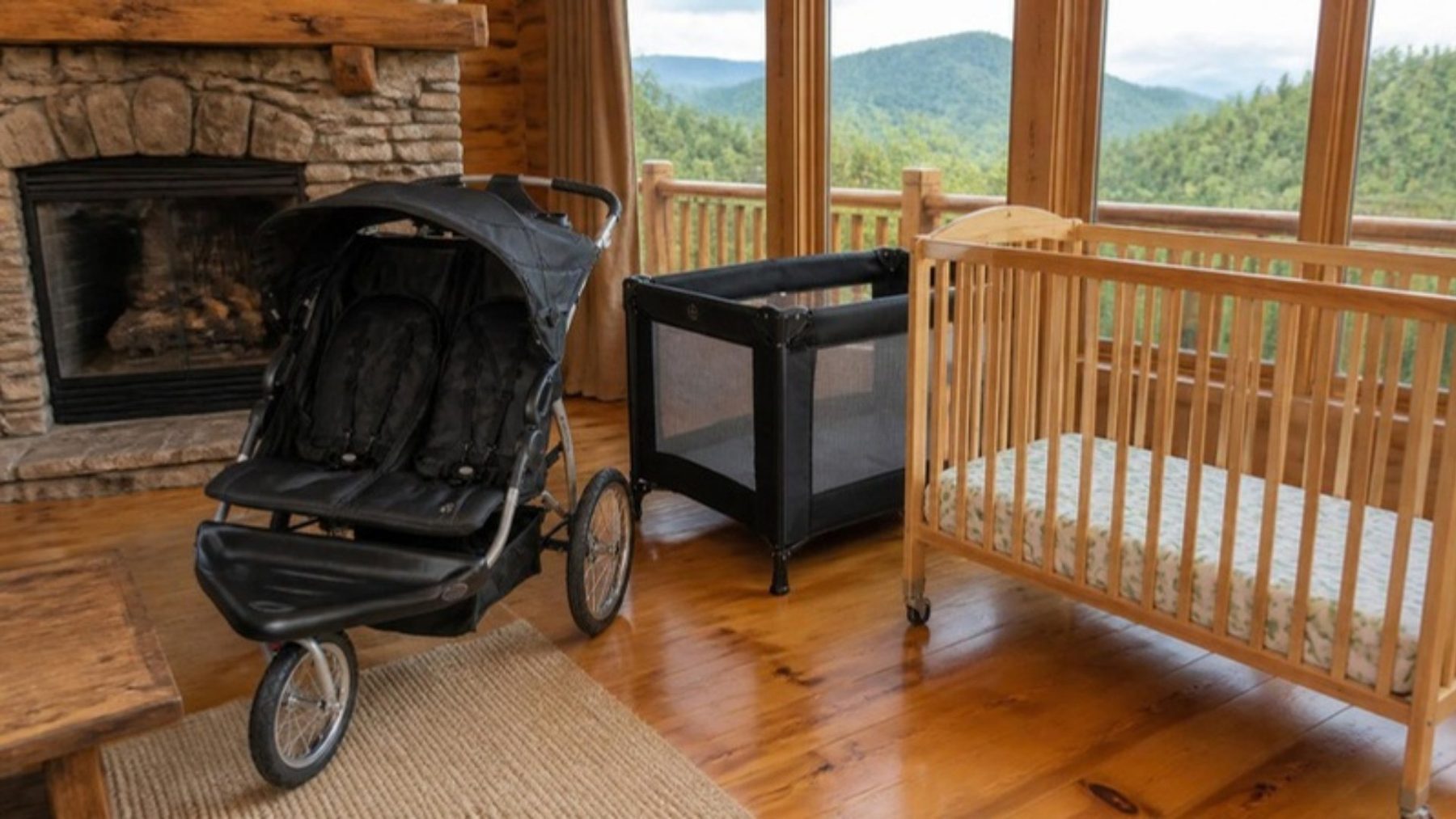 Rental baby gear set up in a Smoky Mountains cabin living room: double jogging stroller, black play yard and wooden crib by a stone fireplace with mountain views