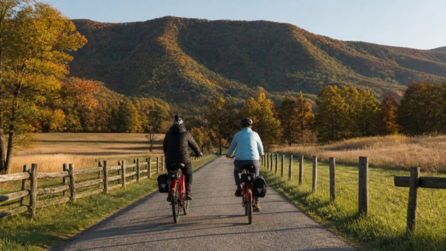 Two riders on red electric bikes with saddlebags ride the Cades Cove Loop road between split-rail fences below an autumn mountain ridge