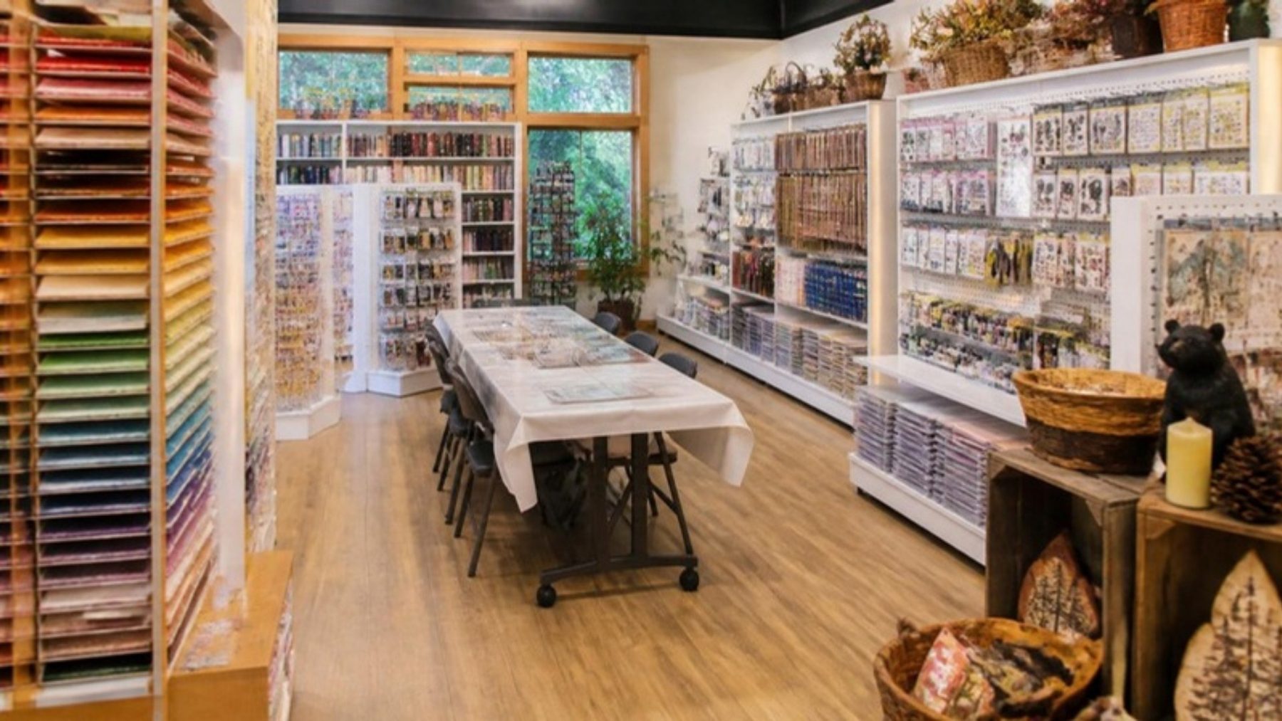 Bright paper crafting store with wall racks of colorful scrapbook paper, shelves of stamps and supplies, and a long class table set for a crop