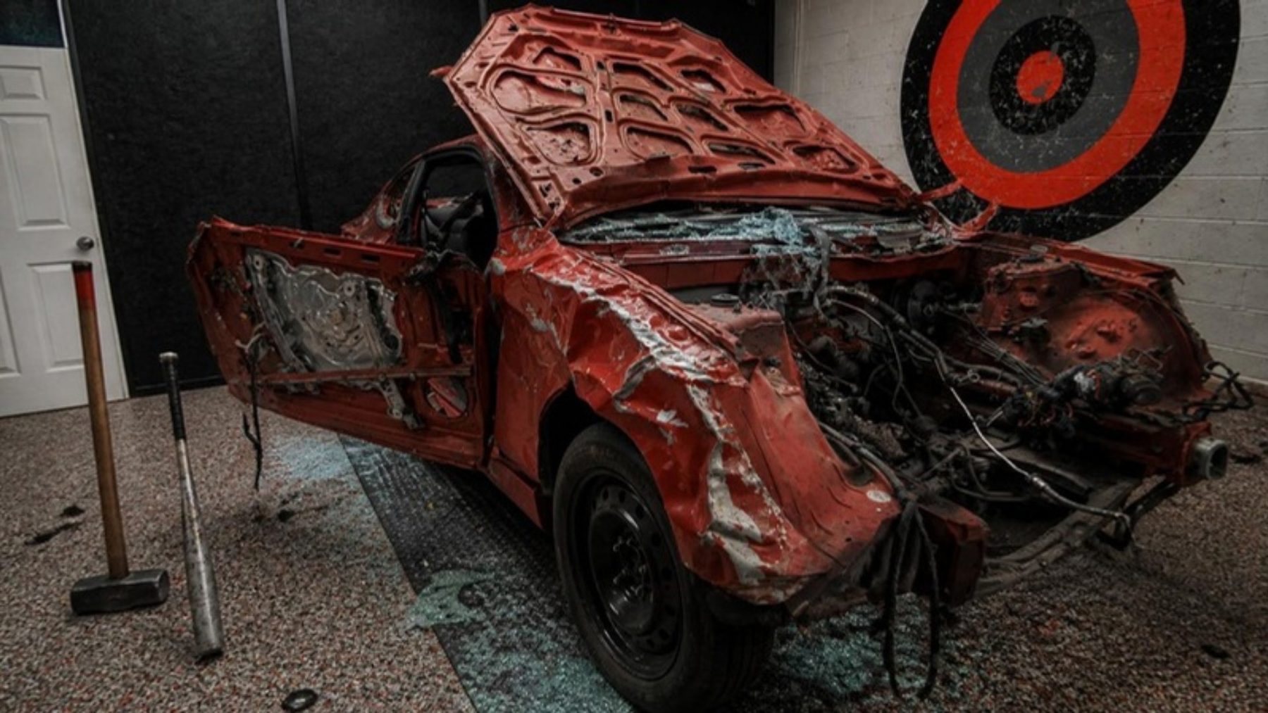 A smashed red sedan inside a dark rage room with a painted target on a white brick wall and a sledgehammer nearby