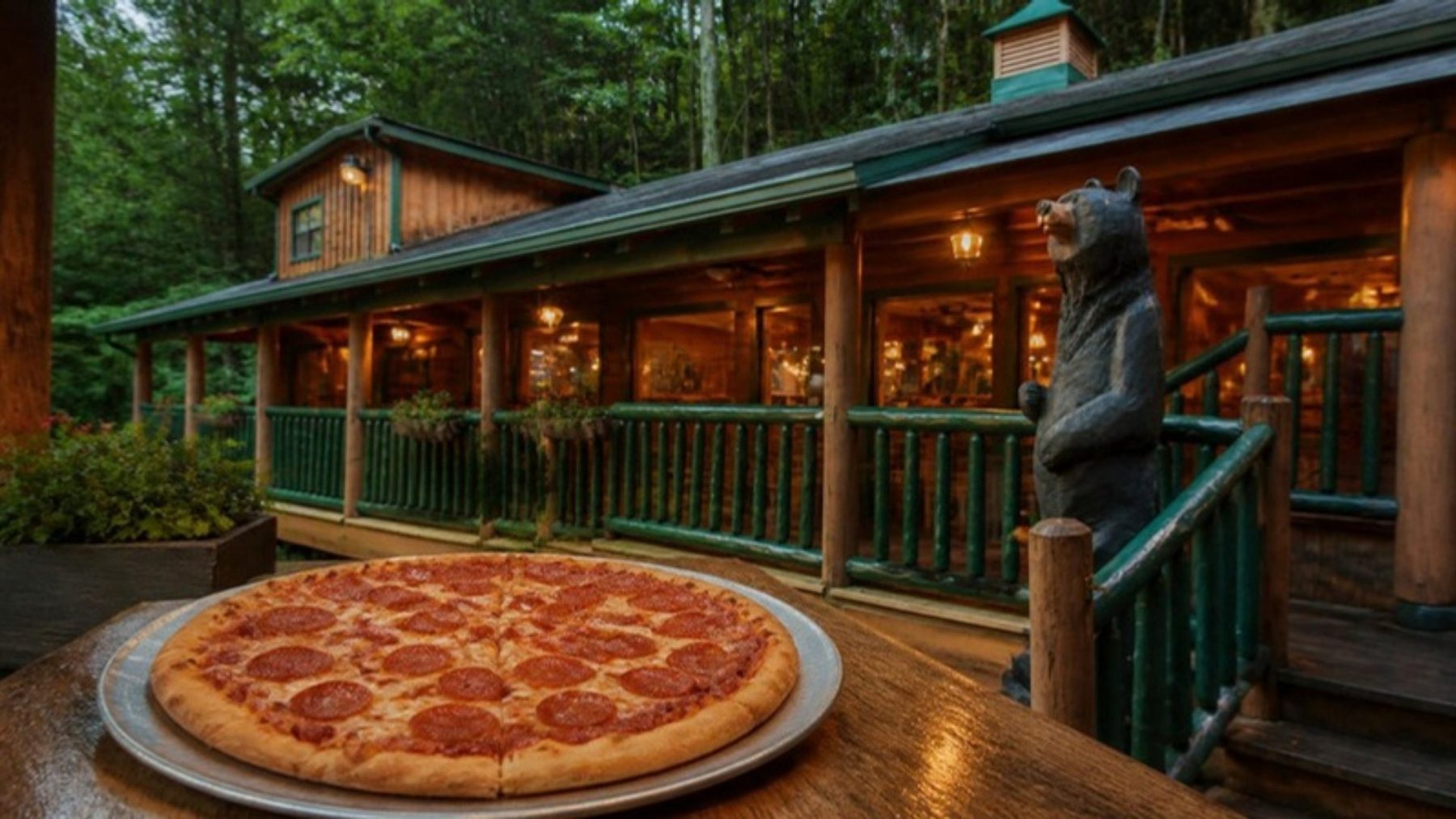 Pepperoni pizza on the porch of a rustic wooded cabin-style pizzeria with green railings and a carved bear statue