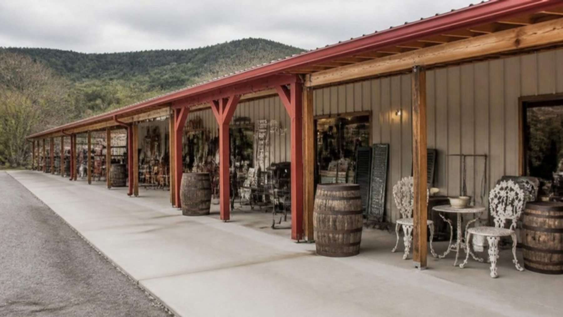 Long metal antique mall building with a red-roofed covered walkway lined with whiskey barrels and white wrought-iron chairs, wooded hills behind