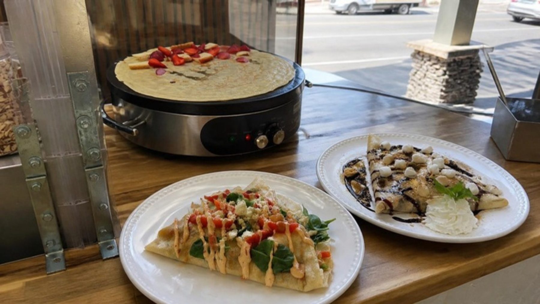 Crepe griddle with a strawberry-topped crepe on a butcher-block counter, with a savory crepe and a dessert crepe plated by a sunny street window