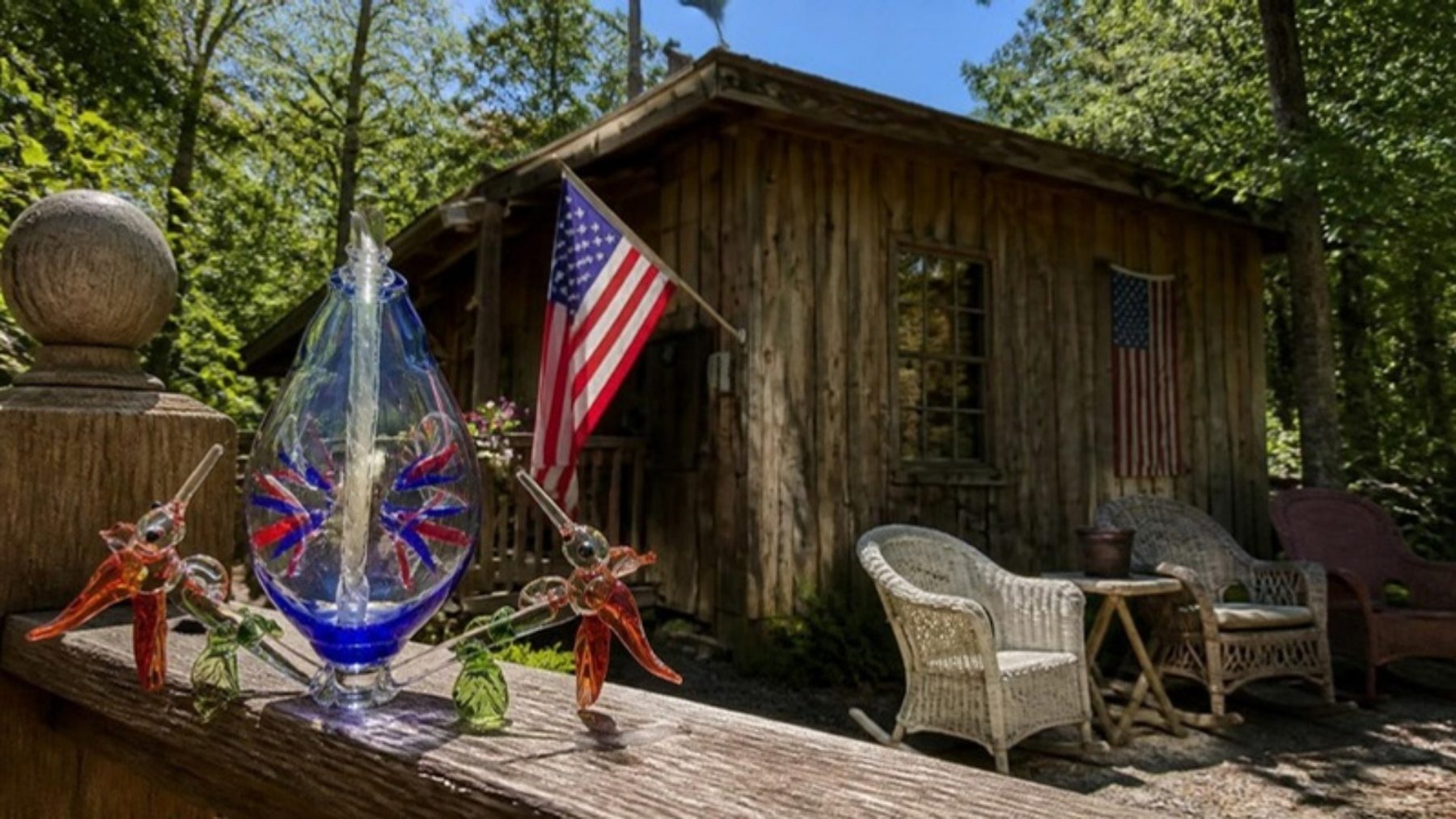 Rustic wooden glassblowing studio among tall trees with an American flag and porch chairs, handmade blue glass vessel with glass hummingbirds on the rail in front