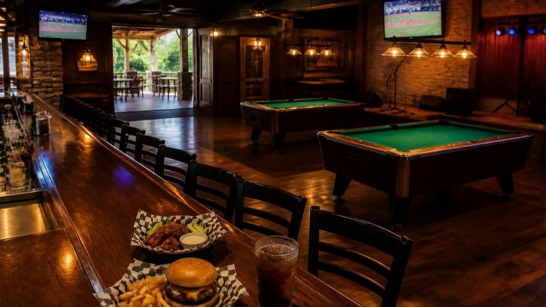 Casual sports bar interior with a long wooden bar, TVs, green-felt pool tables and a basket of bar food in the foreground
