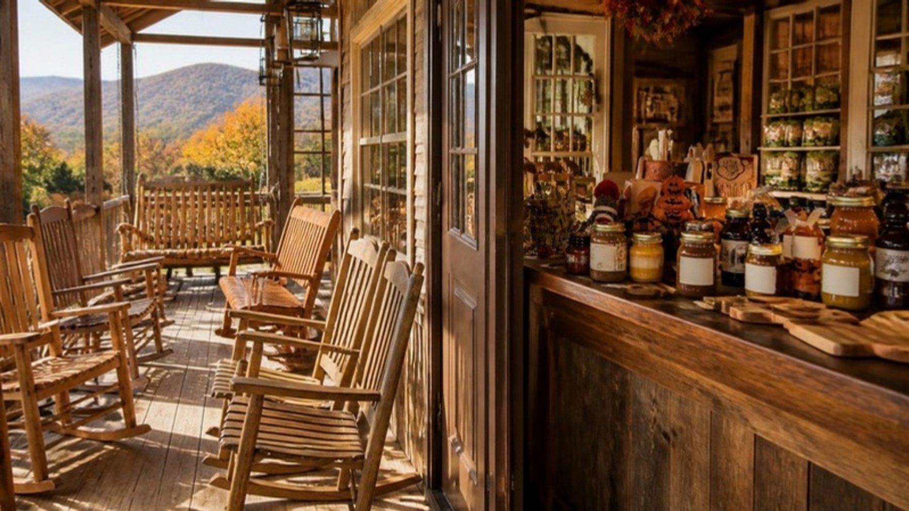 Rustic country store porch with wooden rocking chairs beside a counter of jarred Amish jams, applesauce and syrups, wooded hills behind