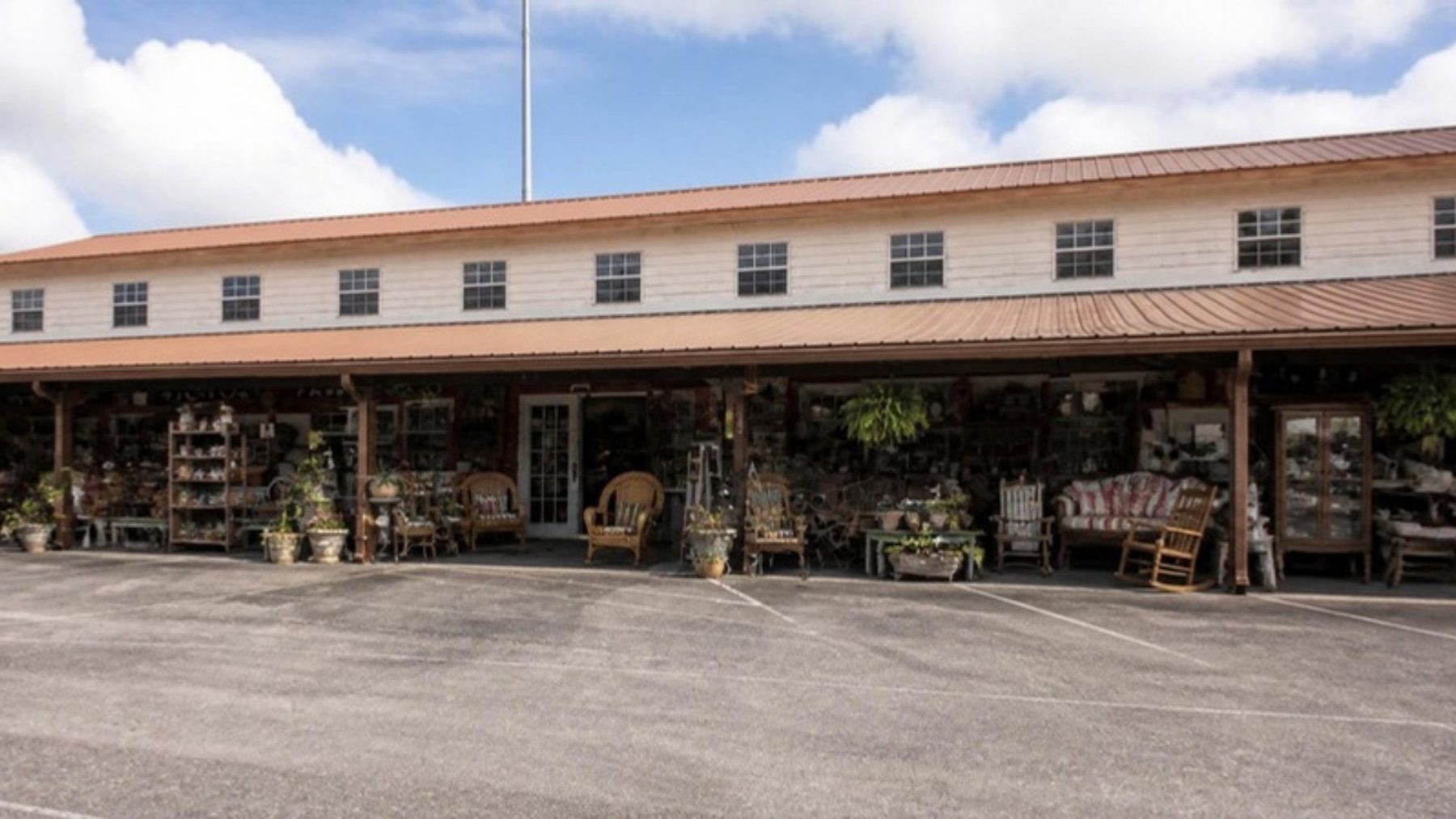 Long, low two-story antique mall with a covered front porch lined with vintage wares, seen across a paved parking lot under a partly cloudy sky