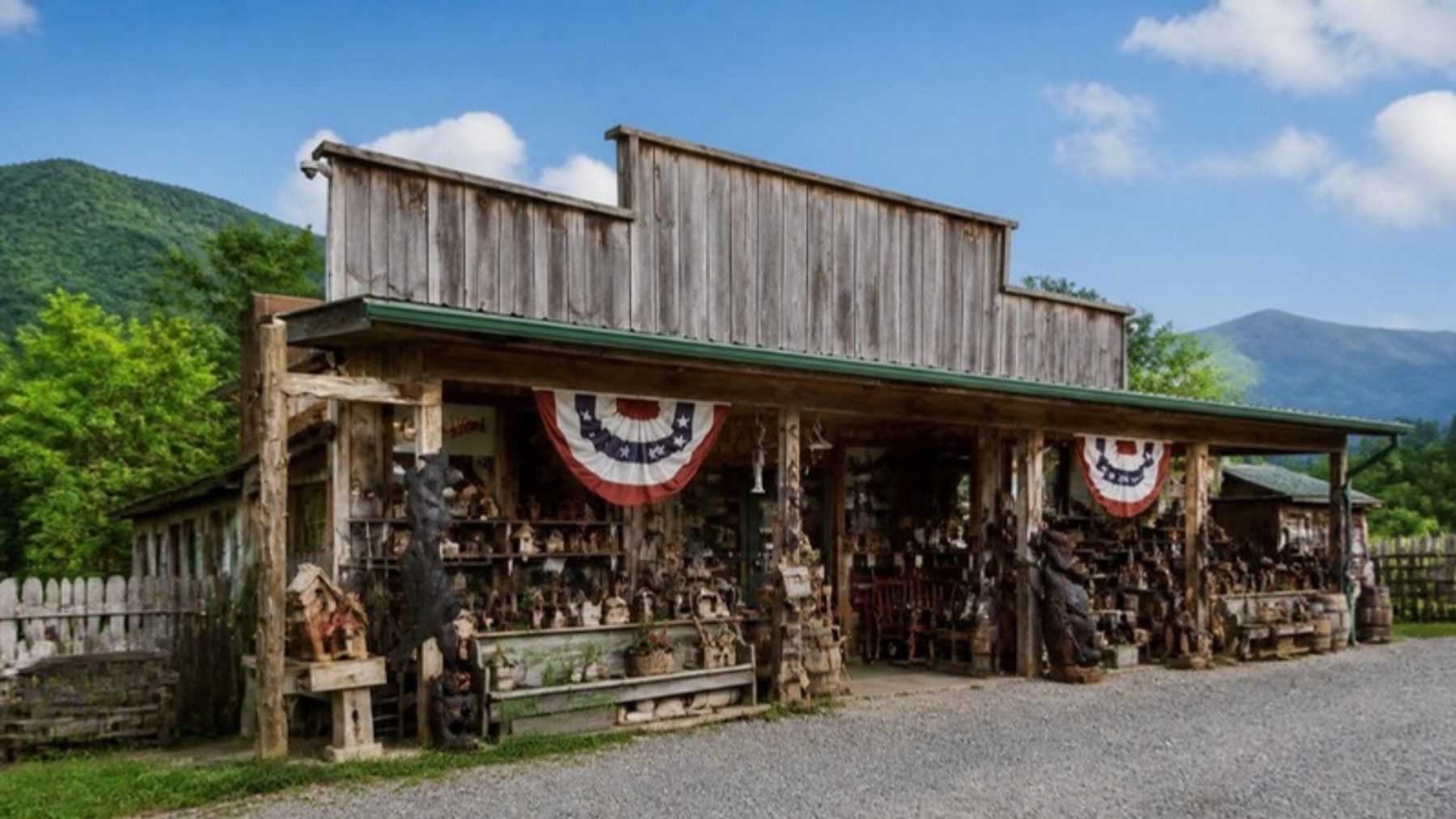 Weathered barnwood country store with a covered porch, patriotic bunting and a porch full of carved wooden bears and rustic wares, green mountains behind