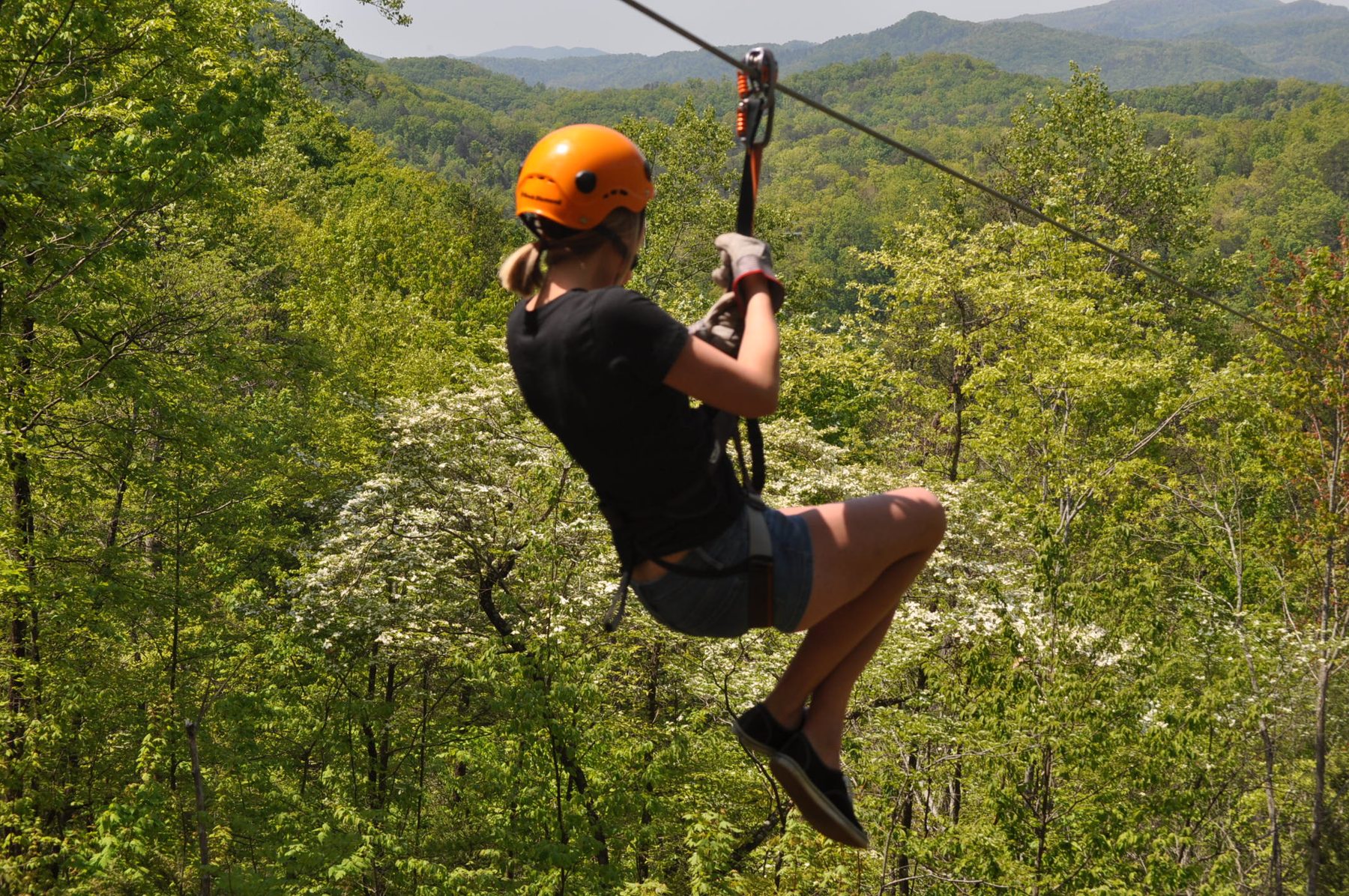 Rider in an orange helmet zipping over a green forest canopy with Smoky Mountain ridges beyond